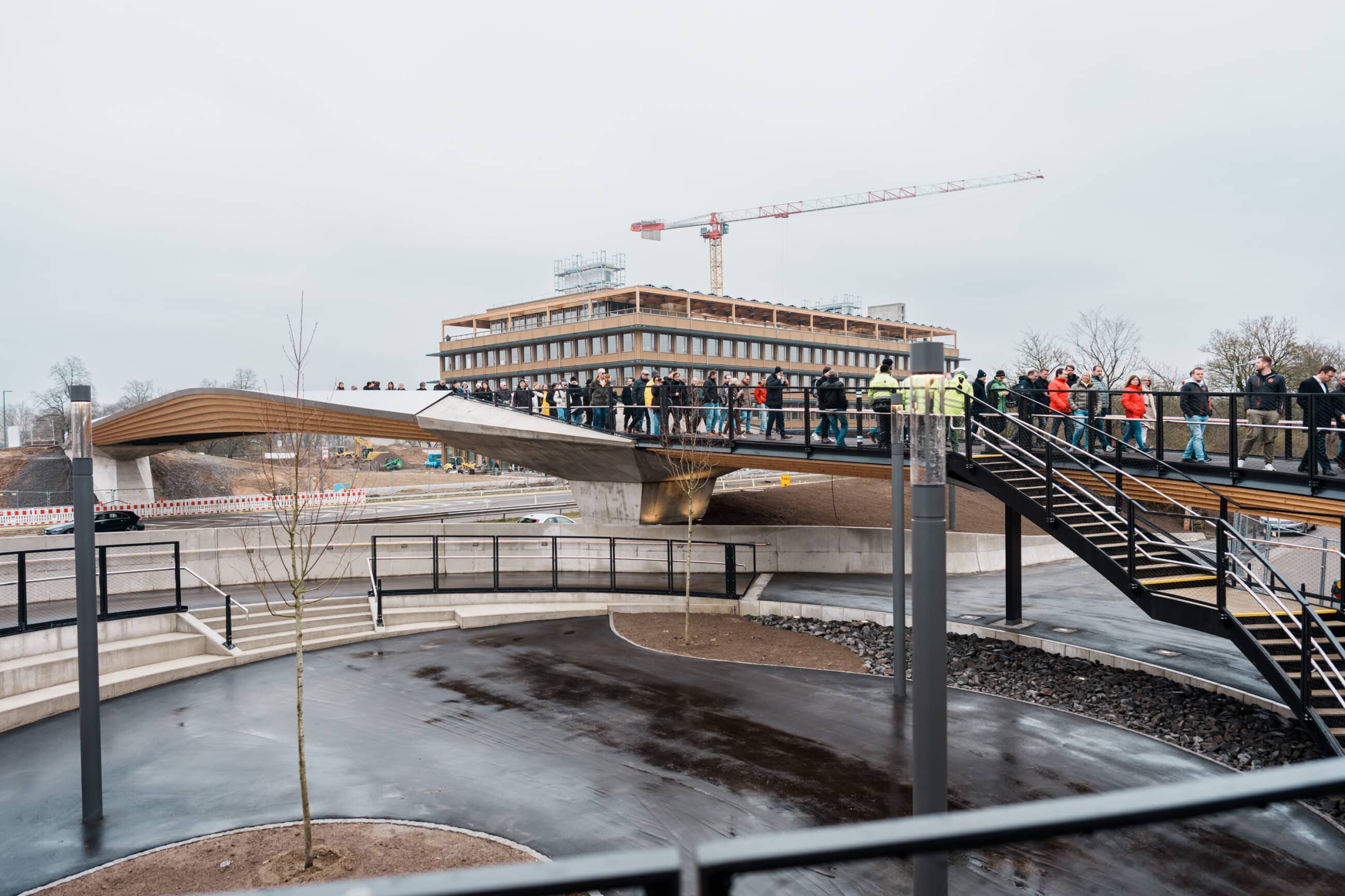 KI-basierte Bildbeschreibung | Eine Gruppe von Menschen steht und geht tagsüber über eine moderne Fußgängerbrücke; im Hintergrund sind ein Baukran und ein Gebäude zu sehen.