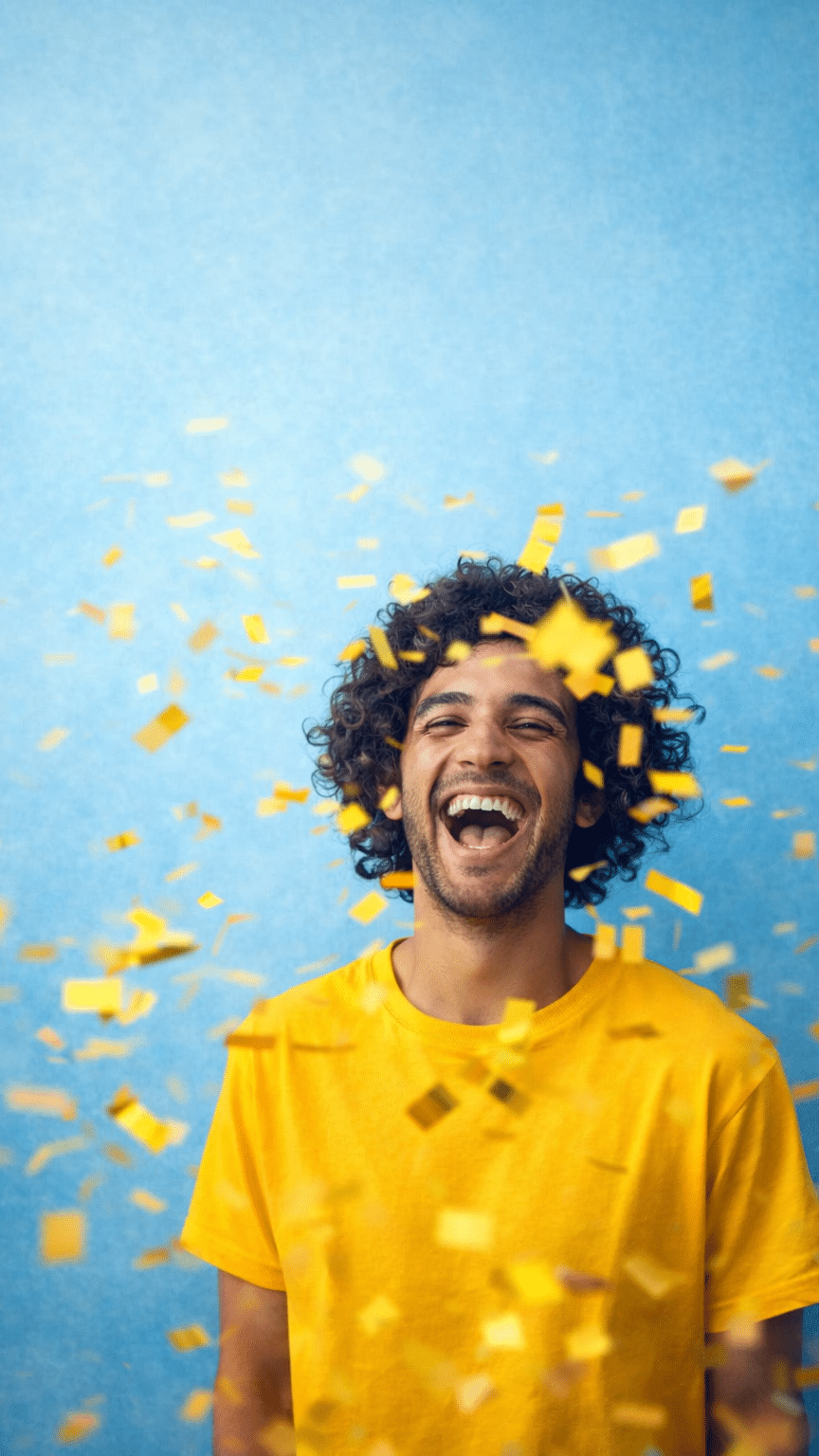 KI-basierte Bildbeschreibung | Ein Mann in einem gelben Hemd steht vor einem blauen Hintergrund, lächelt und lacht, während um ihn herum gelbes Konfetti fällt.